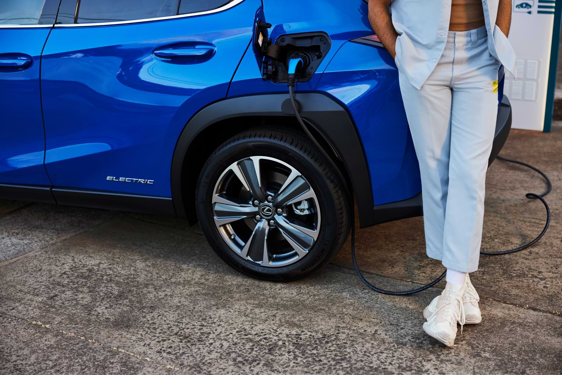 A UX 300e sits charging. The legs of a man leaning against the car as also pictured as he stands next to the charging plug.